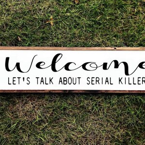 May include: A rectangular wooden sign with a white background and a brown frame. The sign reads "Welcome" in a cursive font, with "LET'S TALK ABOUT SERIAL KILLERS" in a sans-serif font below.