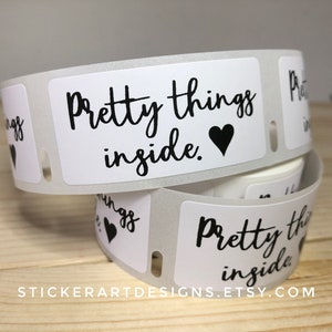 May include: Rolls of white labels with the text "Pretty things inside" in black script, accompanied by a black heart. The labels are designed for packaging and are stacked on top of each other.