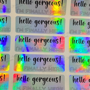 May include: A sheet of holographic stickers with the text "hello gorgeous! I'm finally here" repeated on each sticker. The stickers are iridescent and have a rainbow effect.