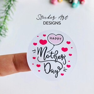 以下が含まれることがあります： ピンクのハート型の風船に黒文字で「Happy」と書かれた白い丸いステッカー。ステッカーには、黒の筆記体で「Mother's Day」と書いてあり、その周りをピンクのハートが囲んでいます。
