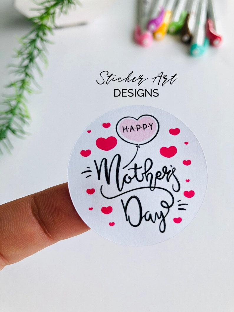 以下が含まれることがあります： ピンクのハートと黒い筆記体で「Happy Mother's Day」と書かれた白い丸いステッカー。ピンクの風船に「H.A.P.P.Y」と書かれており、テキストの上に浮かんでいます。