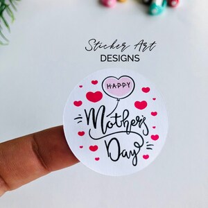 以下が含まれることがあります： ピンクのハート型の風船に「H-A-P-P-Y」と書かれた白い丸いステッカー。黒い筆記体で「母の日」と書かれています。ステッカーは小さなピンクのハートで囲まれています。
