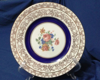 May include: A vintage decorative plate with a floral design. The plate features a white center with a bouquet of pink, orange, and blue flowers, surrounded by a dark blue ring. The outer rim is white with a gold floral pattern.