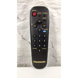 Puede incluir: Mando a distancia de TV Panasonic negro con botones rojos, azules y blancos. El mando tiene botones para encendido, silencio, canal, volumen y los números 0-9. La marca "Panasonic" está impresa en dorado en la parte inferior.