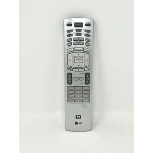 Op de afbeelding: Zilveren LG tv-afstandsbediening met talrijke knoppen voor stroom, volume en kanaalselectie. Voorzien van een TV GUIDE-knop en een numeriek toetsenbord. Het LG-logo is onderaan zichtbaar.