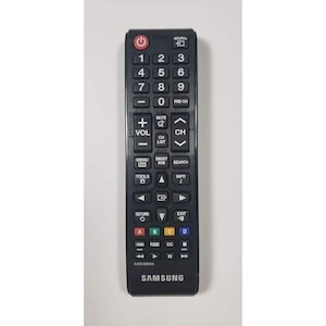 Op de afbeelding: Zwarte Samsung tv-afstandsbediening met talrijke knoppen, waaronder aan/uit, volume, kanaal en menu-opties. Voorzien van een numeriek toetsenbord en knoppen voor smart hub en zoeken. Model AAS9-00854A.
