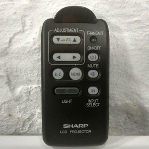 May include: A black Sharp LCD projector remote control. It features buttons for adjustment, volume, transmit, on/off, mute, menu, light, and input select. The remote is rectangular with rounded edges.