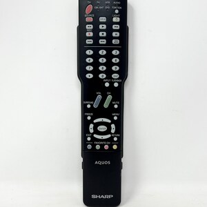 Puede incluir: Un mando a distancia negro Sharp Aquos que presenta un teclado numérico, botones de reproducción, controles de volumen y canal, un anillo de navegación central con botón de entrada y varias teclas de función con texto en blanco.