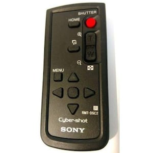 May include: Black Sony Cybershot remote control with various buttons, including "SHUTTER" with a red button, "HOME", "MENU", and directional controls. The remote also features zoom and other function buttons. The model number is RMT-DSC2.