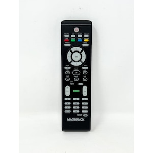 Op de afbeelding: Een zwarte Magnavox afstandsbediening met talrijke knoppen, waaronder aan/uit, bron en navigatie. De afstandsbediening heeft gekleurde knoppen voor functies als rood, groen, blauw en geel. De merknaam "MAGNAVOX" staat onderaan afgedrukt.