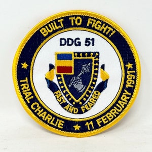 May include: A white, round patch with a blue border and gold stitching. The patch features a shield with a yellow and red flag, a hand holding a spear, and the text "BUILT TO FIGHT! DDG 51 FAST AND FEARED TRIAL CHARLIE 11 FEBRUARY 1991*"