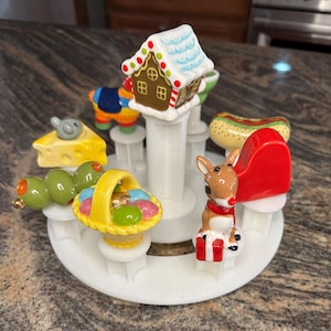 May include: A white ceramic carousel featuring miniature food and holiday-themed figurines. Includes a gingerbread house, hot dog, olives, cheese with a mouse, Easter basket, and a reindeer with a red hat. The carousel is on a brown and gray speckled surface.