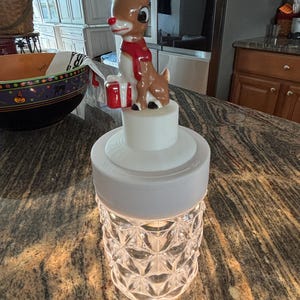 May include: A decorative Christmas-themed night light featuring a Rudolph the Red-Nosed Reindeer figurine atop a clear, diamond-patterned glass base. The figurine is brown with a red scarf and nose, holding a red gift. The night light is illuminated.