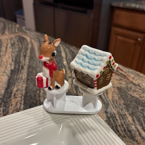 May include: A Christmas-themed serving dish with two decorative bottle stoppers. One stopper features a reindeer with a red scarf and a gift, the other a gingerbread house with white frosting and colorful accents. The stoppers are connected by a white base.