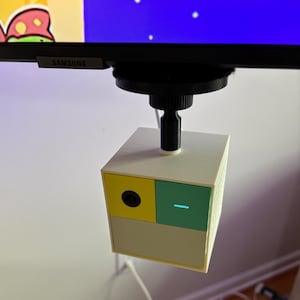 May include: A small, cube-shaped device with a camera lens and a light, mounted below a monitor. The device has a yellow and green panel on the front, with a light blue indicator. The device is attached to a black mount.