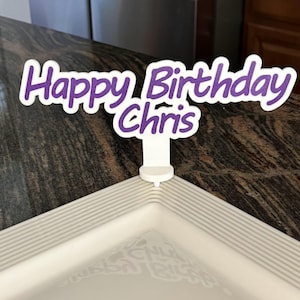 May include: Purple and white Happy Birthday Chris cake topper. The topper is on a white countertop with a black and white patterned surface.