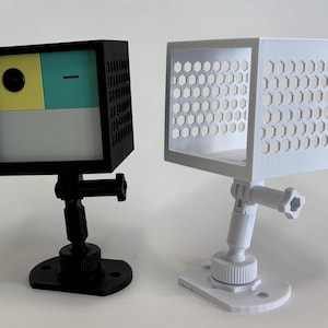 May include: Two cube-shaped camera housings, one black and one white, each mounted on an adjustable stand. The black housing has a color-blocked face with yellow, teal, and white sections. The white housing features a honeycomb pattern.