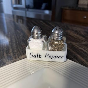 May include: White ceramic salt and pepper shaker holder with the words "Salt Pepper" written on the front. The holder contains two glass shakers, one filled with salt and the other with pepper.