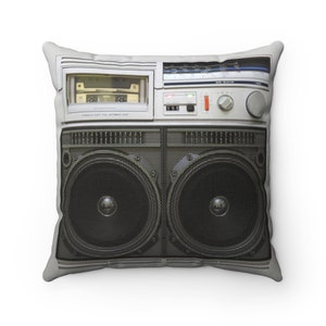 Peut inclure: Un coussin en forme de boombox blanc et noir avec un lecteur de cassette et deux grands haut-parleurs. Le coussin a un fond gris.