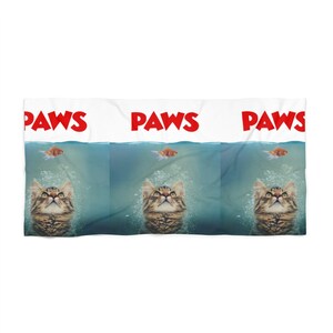 May include: A white beach towel with the word "PAWS" repeated three times in red letters. The towel features a cartoon image of a cat with a surprised expression, looking up at a goldfish swimming above the waterline.