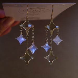 May include: A pair of gold dangle earrings with multiple clear crystal and iridescent moonstone star-shaped charms.