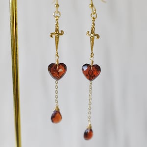 May include: A pair of gold dangle earrings with a dagger charm, a heart-shaped red gemstone, and a teardrop-shaped red gemstone.