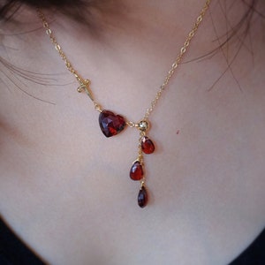 May include: A gold-colored necklace featuring a heart-shaped red gemstone and three teardrop-shaped red gemstones. The necklace has a delicate chain and a small sword charm. The gemstones are faceted, catching the light.