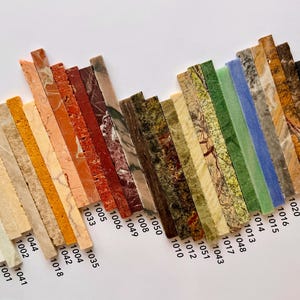 K&ouml;nnte beinhalten: Eine Sammlung rechteckiger Steinproben in verschiedenen Farben und Mustern, diagonal angeordnet. Die Proben umfassen Farbt&ouml;ne von Beige, Orange, Rot, Gr&uuml;n und Blau mit sichtbaren Texturen und Maserungen. Jede Probe ist mit einer Nummer versehen.