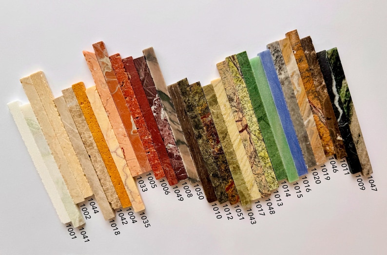 K&ouml;nnte beinhalten: Eine Sammlung rechteckiger Steinproben in verschiedenen Farben und Mustern, diagonal angeordnet. Die Proben umfassen Farbt&ouml;ne von Beige, Orange, Rot, Gr&uuml;n und Blau mit sichtbaren Texturen und Maserungen. Jede Probe ist mit einer Nummer versehen.