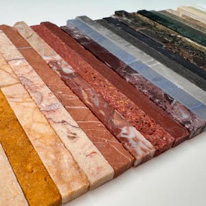 K&ouml;nnte beinhalten: Eine Sammlung rechteckiger Steinproben in verschiedenen Farben und Mustern. Die Proben umfassen Farbt&ouml;ne von Beige, Orange, Rot, Braun, Grau und Gr&uuml;n. Die Texturen variieren von glatt bis leicht rau und zeigen die nat&uuml;rlichen Variationen im Stein.