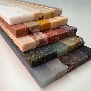 K&ouml;nnte beinhalten: Eine Sammlung rechteckiger Steinfliesen in verschiedenen Farben, darunter Wei&szlig;, Beige, Rot, Braun, Gr&uuml;n und Schwarz. Die Fliesen sind in einer gestuften Anordnung angeordnet, die die unterschiedlichen Texturen und Farbt&ouml;ne des Natursteins zeigt.