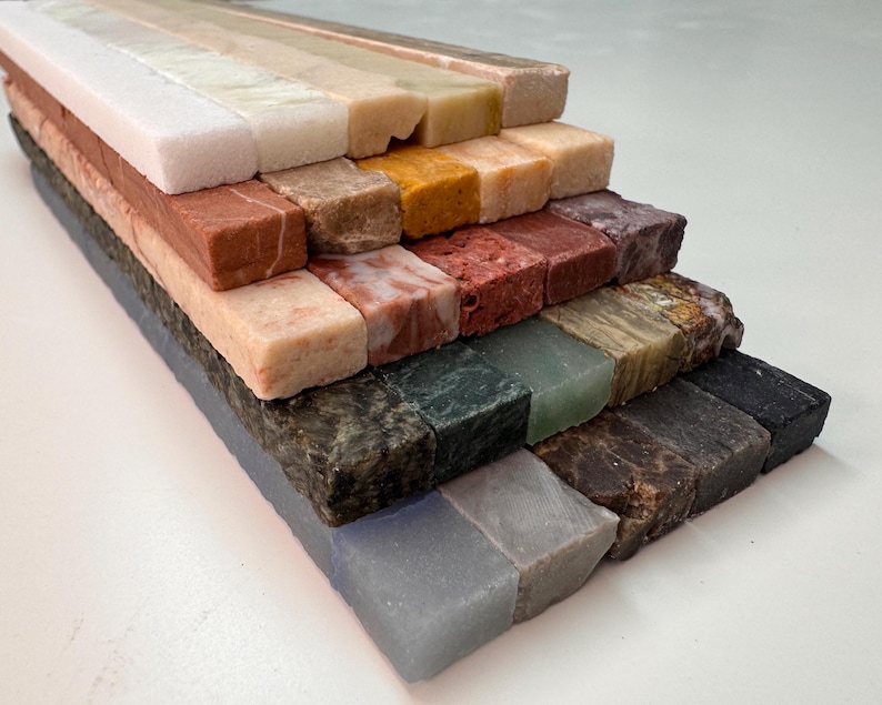 K&ouml;nnte beinhalten: Eine Sammlung rechteckiger Steinfliesen in verschiedenen Farben, darunter Wei&szlig;, Beige, Rot, Braun, Gr&uuml;n und Schwarz. Die Fliesen sind in einer gestuften Anordnung angeordnet, die die unterschiedlichen Texturen und Farbt&ouml;ne des Natursteins zeigt.