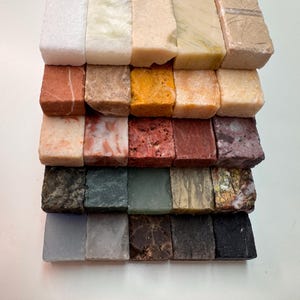 K&ouml;nnte beinhalten: Eine Sammlung kleiner, rechteckiger Steinproben in verschiedenen Farben und Texturen. Die Proben umfassen Farbt&ouml;ne von Wei&szlig;, Beige, Rot, Braun, Gelb, Gr&uuml;n und Schwarz. Jedes St&uuml;ck hat ein einzigartiges Muster und eine einzigartige Oberfl&auml;che.