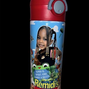 May include: A cylindrical water bottle with a red lid and a light blue body featuring a photographic portrait of a young child with braided hair. The design includes cartoon characters and the text: REMIDI IS... SMART. KIND. FUNNY & PRETTY REMIDI'S STORY.