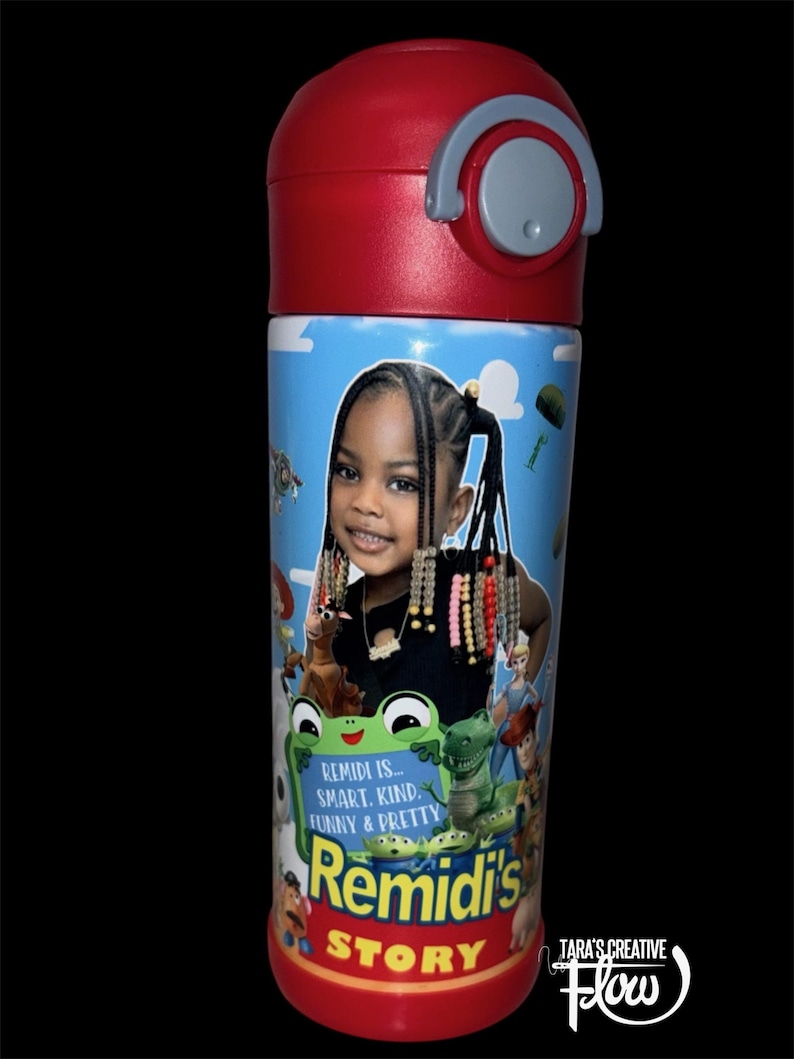 May include: A cylindrical water bottle with a red lid and a light blue body featuring a photographic portrait of a young child with braided hair. The design includes cartoon characters and the text: REMIDI IS... SMART. KIND. FUNNY & PRETTY REMIDI'S STORY.