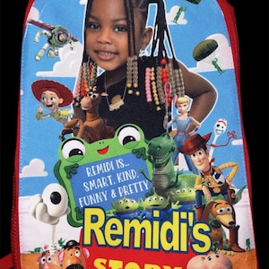 May include: A blue backpack featuring a photographic portrait of a young child with braided hair, surrounded by various animated characters. The text reads: REMIDI IS... SMART, KIND, FUNNY & PRETTY, Remidi's STORY. The bag has a red zipper border.