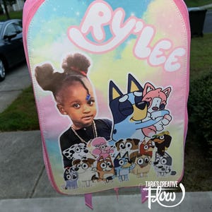 May include: A pink backpack featuring a pastel tie-dye background with the name RY'LEE printed in white at the top. The front displays a photographic portrait of a child alongside a collection of cartoon dog characters from the Bluey series.