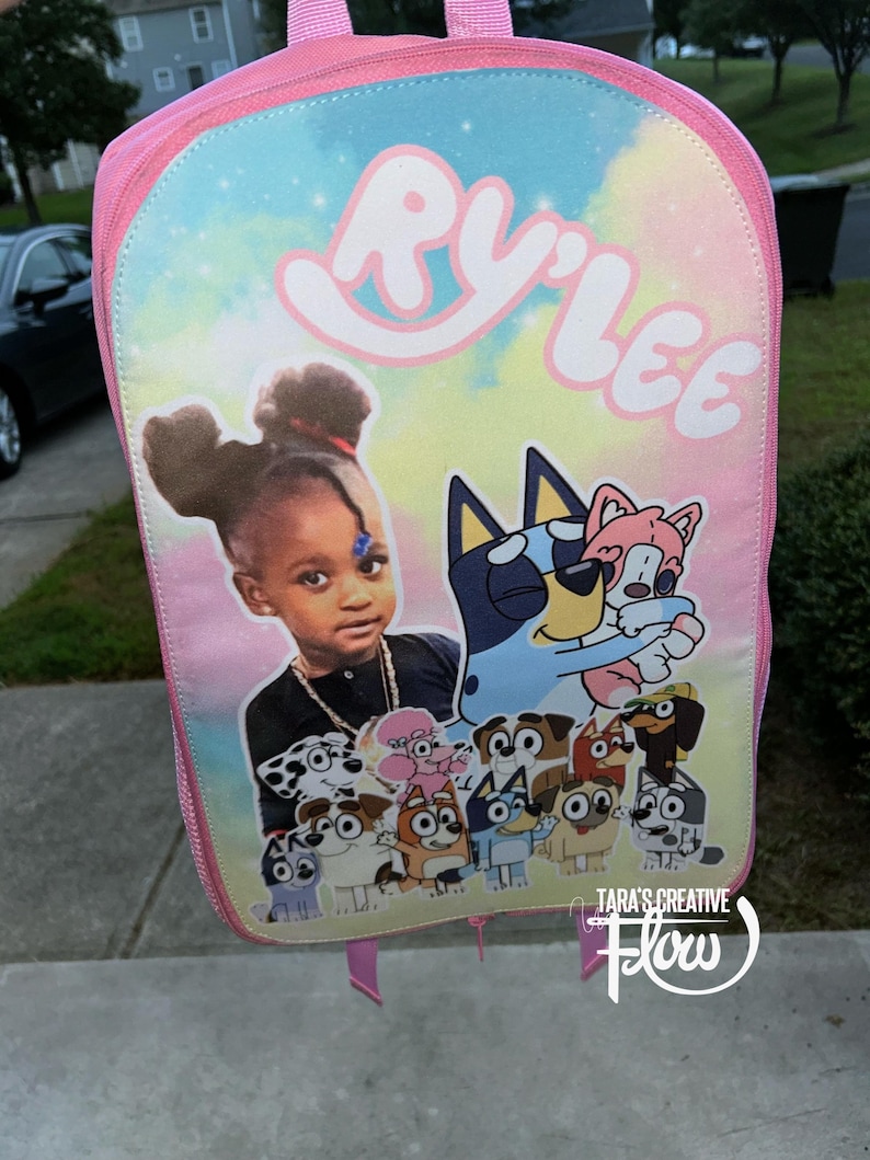 May include: A pink backpack featuring a pastel tie-dye background with the name RY'LEE printed in white at the top. The front displays a photographic portrait of a child alongside a collection of cartoon dog characters from the Bluey series.