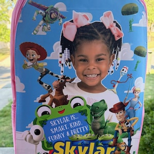 May include: A light blue backpack featuring a custom graphic print of a young child surrounded by various animated toy characters. The design includes the text Skylar is... Smart, Kind, Funny & Pretty and Skylar Story in bold yellow lettering.