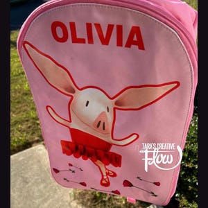 May include: A light pink backpack featuring a graphic of a cartoon pig wearing a red tutu and ballet slippers. The name OLIVIA is printed in bold red capital letters at the top. Small red rose illustrations are scattered near the bottom.