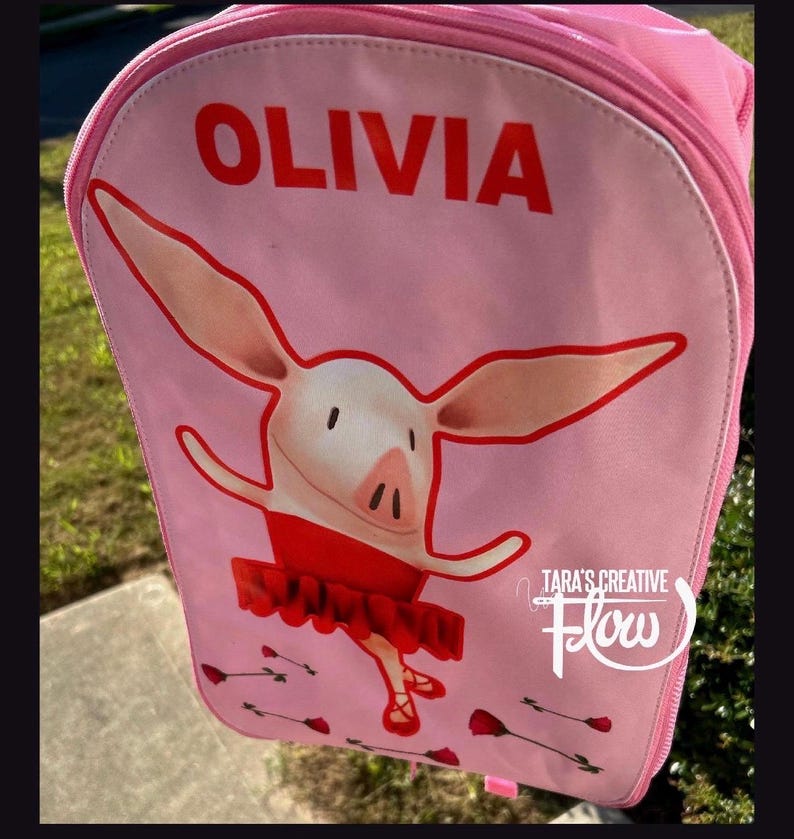 May include: A light pink backpack featuring a graphic of a cartoon pig wearing a red tutu and ballet slippers. The name OLIVIA is printed in bold red capital letters at the top. Small red rose illustrations are scattered near the bottom.