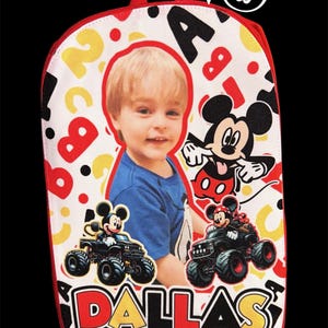 May include: A white backpack featuring a red trim and a graphic print of a child, Mickey Mouse, and two monster trucks. The design includes scattered red and yellow letters and numbers, with the name DALLAS printed in large, bold, multi-colored letters.