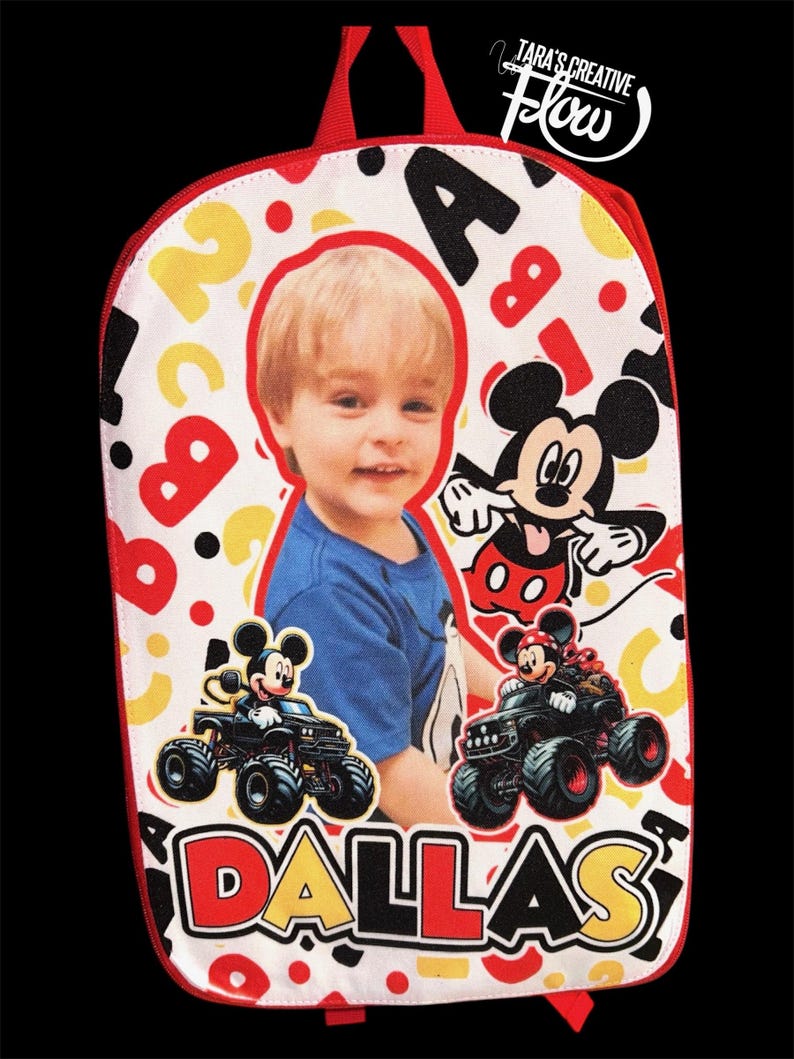 May include: A white backpack featuring a red trim and a graphic print of a child, Mickey Mouse, and two monster trucks. The design includes scattered red and yellow letters and numbers, with the name DALLAS printed in large, bold, multi-colored letters.
