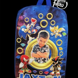 May include: A blue backpack featuring a galaxy print, Sonic the Hedgehog characters, and a central circular frame containing a photograph of a young child. The name JAXTON is printed in large blue letters at the bottom, surrounded by gold rings.
