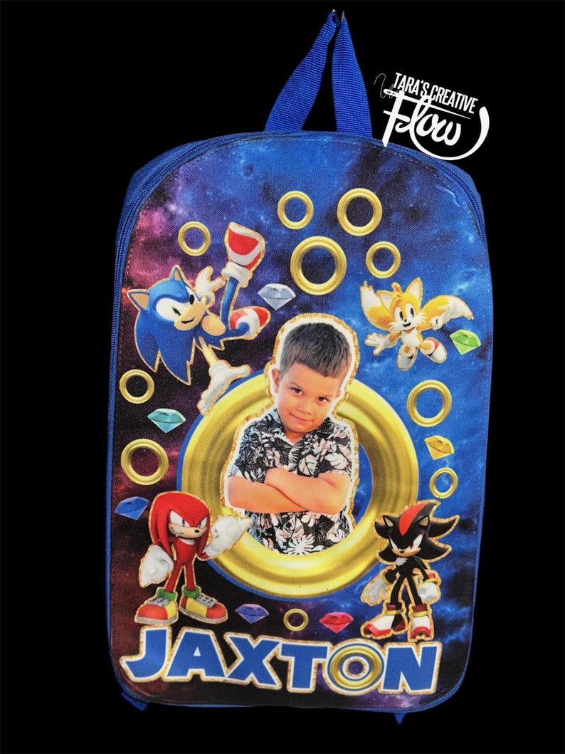May include: A blue backpack featuring a galaxy print, Sonic the Hedgehog characters, and a central circular frame containing a photograph of a young child. The name JAXTON is printed in large blue letters at the bottom, surrounded by gold rings.