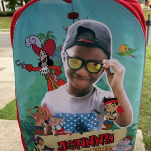 May include: A red backpack featuring a custom graphic of a child wearing a grey cap and yellow-tinted sunglasses, surrounded by cartoon characters from Never Land Pirates. The text JOHNNY and NEVER LAND PIRATES is printed at the bottom of the image.