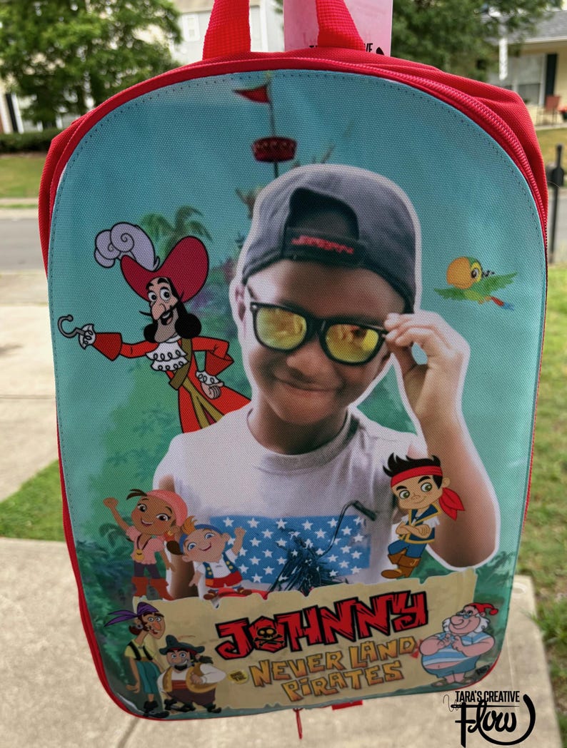 May include: A red backpack featuring a custom graphic of a child wearing a grey cap and yellow-tinted sunglasses, surrounded by cartoon characters from Never Land Pirates. The text JOHNNY and NEVER LAND PIRATES is printed at the bottom of the image.