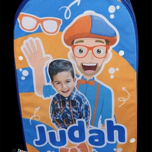 May include: A blue and orange backpack featuring a graphic print of a cartoon character wearing an orange hat and glasses, alongside a photograph of a young child. The name Judah is printed in large blue letters, with a small orange bow tie below.