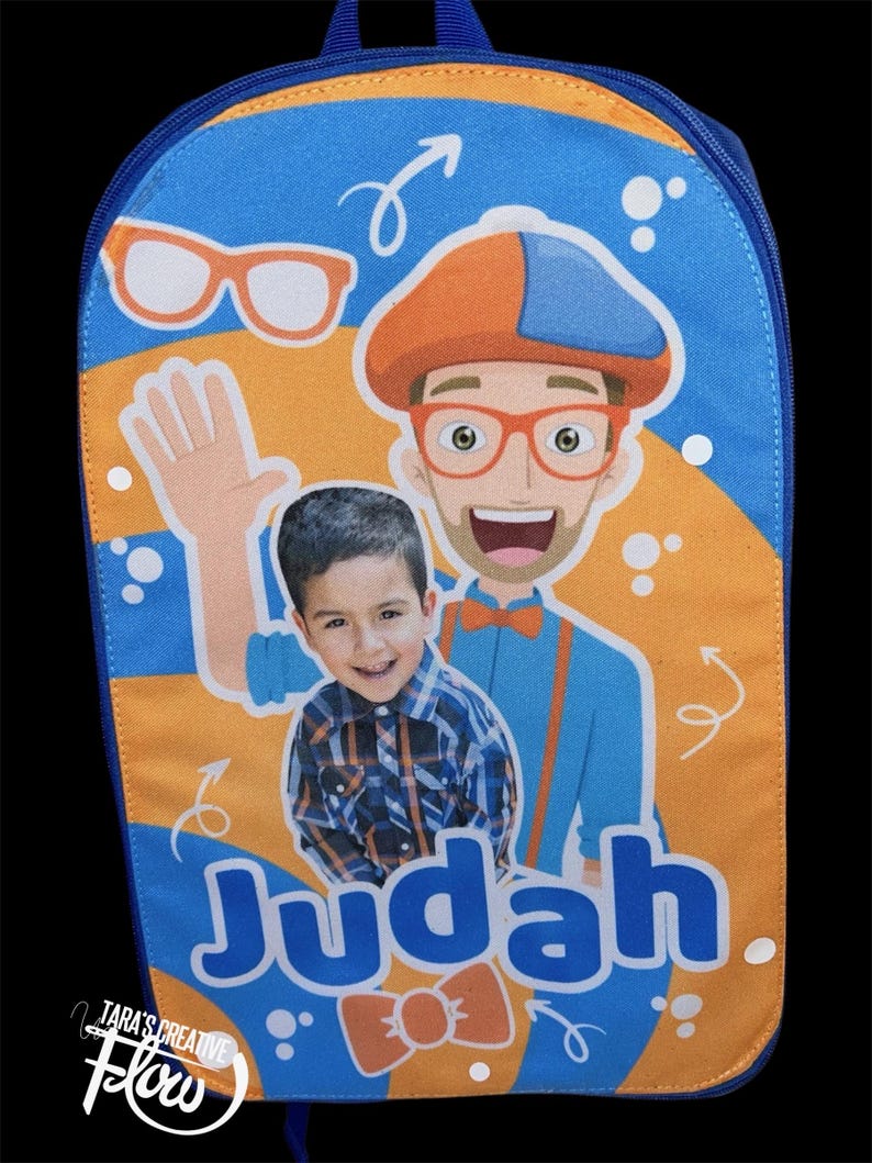 May include: A blue and orange backpack featuring a graphic print of a cartoon character wearing an orange hat and glasses, alongside a photograph of a young child. The name Judah is printed in large blue letters, with a small orange bow tie below.