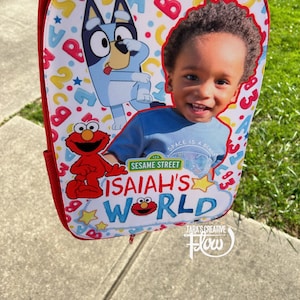 May include: A red and white backpack with a Sesame Street theme. The backpack features a blue dog character, Elmo, and a child's face. The text "ISAIAH'S WORLD" is printed on the backpack.